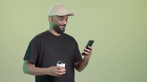 التأكد من مطابقة الطلب قبل تسليمه عبر الموبايل, استخدام التكنولوجيا الحديثة في الحياة العملية، توصيل معتمد ومضمون, توصيل المشروبات الساخنة، بورتريه لمندوب عربي خليجي سعودي مبتسم يرتدي الزي الكاجوال و قبعة بيج يحمل كوب ساخن و الهاتف المحمول, خلفية خضراء