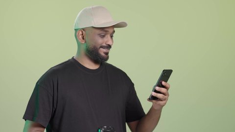 التحقق من تفاصيل الطلب أثناء التوصيل عبر الهاتف المحمول, توصيل المشروبات الساخنة، بورتريه لمندوب عربي خليجي سعودي مبتسم يرتدي الزي الكاجوال و قبعة بيج يحمل كوب ساخن و الجوال, خلفية خضراء