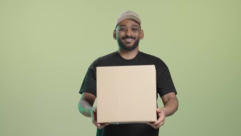 توصيل معتمد ومضمون, ايماءات السعادة و السرور, بورتريه لمندوب عربي خليجي سعودي يرتدي الزي الكاجوال و قبعة بيج يحمل صندوق كرتوني و يسلمه بابتسامة, خلفية خضراء