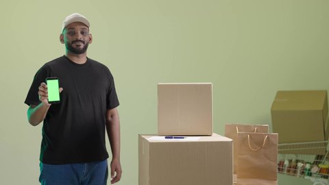 استخدام التكنولوجيا الحديثة في الحياة العملية، توصيل معتمد ومضمون, ايماءات السعادة و السرور, بورتريه لمندوب عربي خليجي سعودي مبتسم يرتدي الزي الكاجوال و قبعة بيج يحمل هاتف محمول و يقوم بعرض الشاشة, خلفية خضراء