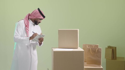 Ensuring the safety of parcels and goods sent, the concept of e-shopping, a reliable shipping and delivery process, a portrait of a Saudi Arabian Gulf representative wearing traditional clothing and a shemagh, writing down and reviewing the order, a green background