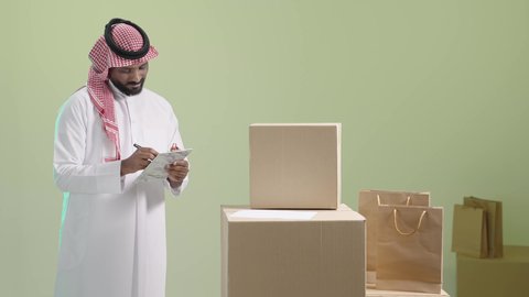 الكراتين المغلقة بمجموعة متنوعة من الأحجام والأشكال، مفهوم التسوق الالكتروني, عملية شحن وتوصيل موثوقة, بورتريه لمندوب عربي خليجي سعودي يرتدي الزي التقليدي والشماغ يقوم بتدوين و مراجعة الطلب, خلفية خضراء