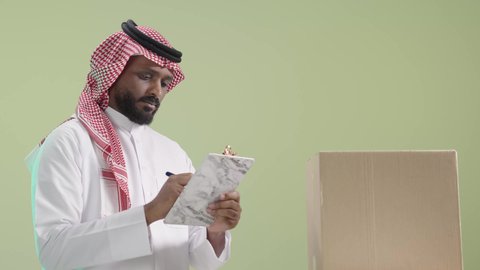 تنظيم قائمة الطلبيات قبل مرحلة التغليف, عملية شحن وتوصيل موثوقة, بورتريه لمندوب عربي خليجي سعودي يرتدي الزي التقليدي والشماغ يقوم بتدوين الطلب, خلفية خضراء