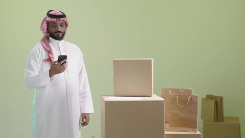 ايماءات الرضا والسرور بتوصيل الطلبات للزبائن، طلب المنتجات عبر تطبيقات الهاتف المحمول، استخدام الأجهزة التقنية الحديثة، بورتريه لرجل عربي خليجي سعودي مبتسم يرتدي الثوب التقليدي والشماغ يحمل الجوال, عملية شحن و توصيل موثوقة, خلفية خضراء