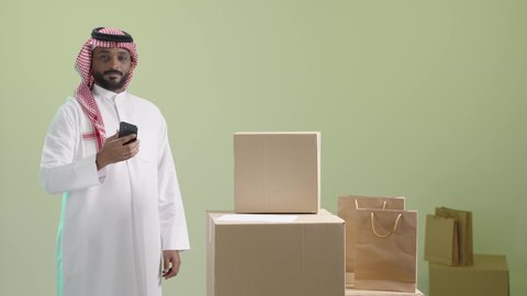 أداء العمل بدقة وإتقان، طلب المنتجات عبر تطبيقات الهاتف المحمول،  استخدام الأجهزة التقنية الحديثة، بورتريه لرجل عربي خليجي سعودي مبتسم يرتدي الثوب التقليدي والشماغ يحمل الجوال, عملية شحن و توصيل موثوقة, خلفية خضراء