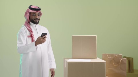 Ordering products via mobile applications, using modern technology devices, portrait of a smiling Saudi Arabian Gulf man wearing traditional thobe and shemagh holding a mobile phone and standing next to cardboard boxes, displaying the mobile screen, green background