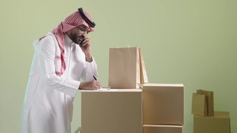خدمة التوصيل المنزلي، ادارة عملية الشحن, بورتريه لرجل عربي خليجي سعودي يرتدي الزي التقليدي والشماغ يقوم بالتواصل مع العميل من خلال الجوال و تدوين الطلب, خلفية خضراء