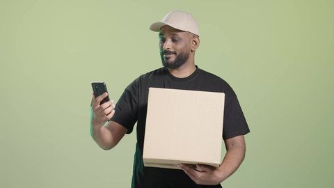 مراجعة الطلب من خلال الجوال, الاستفادة من التكنولوجيا الحديثة بتسهيل العمل، بورتريه لمندوب عربي خليجي سعودي مبتسم يرتدي الزي الكاجوال و قبعه بيج يحمل هاتف محمول و صندوق كرتوني, خلفية خضراء