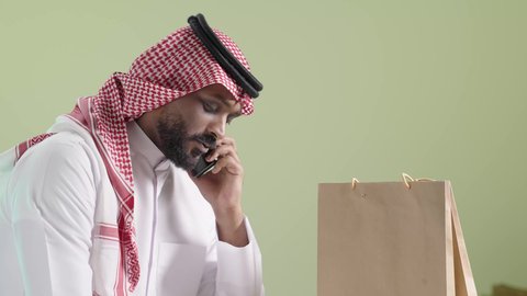 Recording order details while preparing parcels, high quality shipping and delivery service, portrait of a Saudi Arabian Gulf man wearing traditional clothing and a shemagh communicating with the customer via mobile phone and recording the order, green background