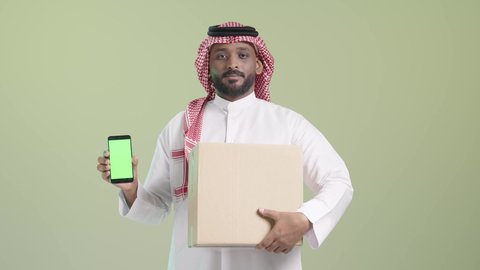 الشراء والتوصيل عبر تطبيقات الهاتف المحمول, عرض شاشة الموبايل، الاستفادة من التكنولوجيا الحديثة بتسهيل العمل، بورتريه لرجل عربي خليجي سعودي مبتسم يرتدي الزي التقليدي والشماغ يحمل الجوال و صندوق كرتوني, عملية شحن و توصيل موثوقة, خلفية خضراء