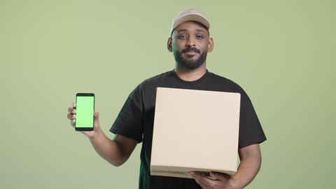 عرض شاشة الموبايل، الاستفادة من التكنولوجيا الحديثة بتسهيل العمل، بورتريه لمندوب عربي خليجي سعودي مبتسم يرتدي الزي الكاجوال و قبعه بيج يحمل هاتف محمول و صندوق كرتوني, خلفية خضراء