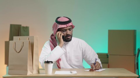 مراجعة فواتير الشحن على مكتب المندوب, بورتريه لرجل عربي خليجي سعودي يرتدي الزي التقليدي والشماغ يقوم بالتواصل مع العميل من خلال الجوال و تدوين الطلب, خلفية ملونة