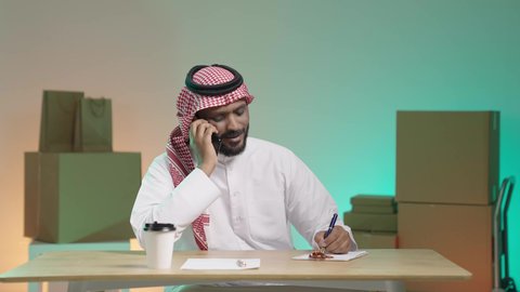 Follow up on delivery orders during a phone call, high quality shipping and delivery service, portrait of a Saudi Arabian Gulf man wearing traditional clothing and a shemagh communicating with the customer via mobile phone and writing down the order, colorful background