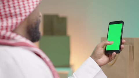استخدام الجوال والتكنولوجيا الحديثة لتسهيل القيام بالمهام اليومية, عرض شاشة الموبايل، بورتريه من الخلف لرجل عربي خليجي سعودي يرتدي الزي التقليدي والشماغ يمسك هاتف محمول و يباشر العمل من خلاله