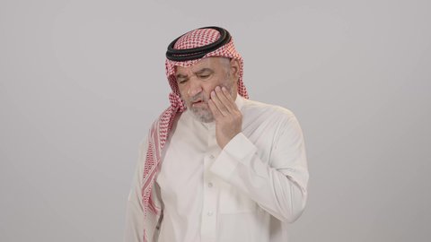 أهمية زيارة الطبيب والحصول على قسط من الراحة, تسوس الأضراس و إلتهاب اللثه، طلب العلاج و المساعدة، بورتريه لرجل مسن عربي خليجي سعودي يرتدي الثوب التقليدي و الشماغ يضع يديه على خده و يغمض عينيه بإيماءات الشعور بألم الأسنان