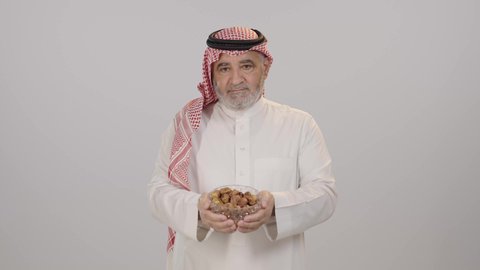 صحن مليء بالتمر, ابتسامة صادقة تعكس روح الكرم, الترحيب بالضيف وتقدير قيم المشاركة والود, بورتريه لرجل مسن عربي خليجي سعودي يرتدي الثوب التقليدي و الشماغ يؤدي واجب الضيافة