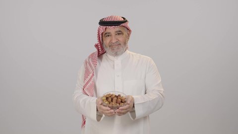 ابتسامة صادقة تعكس روح الكرم, الترحيب بالضيف وتقدير قيم المشاركة والود, بورتريه لرجل مسن عربي خليجي سعودي يرتدي الثوب التقليدي و الشماغ يؤدي واجب الضيافة و يقدم صحن مليء بالتمر