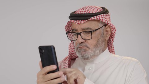 Using modern and advanced technologies, communicating and chatting with family and friends via mobile applications, a portrait of an elderly Saudi Arabian Gulf man wearing traditional thobe and shemagh, wearing glasses, browsing social media sites