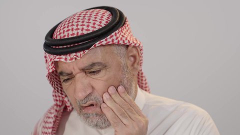 طلب العلاج و المساعدة، بورتريه لرجل مسن عربي خليجي سعودي يرتدي الثوب التقليدي و الشماغ يضع يديه على خده و يغمض عينيه بإيماءات الشعور بألم الأسنان, تسوس الأضراس وإلتهاب اللثه, تعابير الوجع والتألم