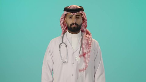 الوقوف باستقامة بايماءات ايدي متقاطعة, الاستعداد لتقديم الخدمات للمرضى بكل كفاءة ،مفهوم الطب والرعاية الصحية، بورتريه لطبيب عربي خليجي سعودي مبتسم يرتدي الثوب التقليدي والشماغ مع المعطف الأبيض يضع سماعة الفحص ينظر إلى الكاميرا بتعابير السعادة والسرور, خلفية زرقاء