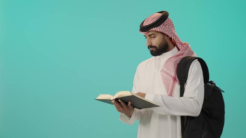 Reading university books, reviewing daily lessons, portrait of a smiling Saudi Arabian Gulf man wearing traditional white thobe, shemagh and backpack holding a university book, preparing for the new school year, blue background