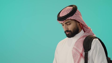 الاستعداد للعام الدراسي الجديد, قراءة الكتب الجامعية، مراجعة الدروس اليومية، بورتريه لرجل عربي خليجي سعودي مبتسم يرتدي الثوب الأبيض التقليدي و الشماغ و حقيبة الظهر يحمل بيده كتاب جامعي, خلفية زرقاء