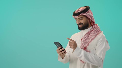 استخدام الجوال والتكنولوجيا الحديثة لتسهيل القيام بالمهام اليومية, بورتريه لشاب عربي سعودي خليجي مبتسم يرتدي الثوب التقليدي والشماغ يستخدم الهاتف المحمول, خلفية زرقاء