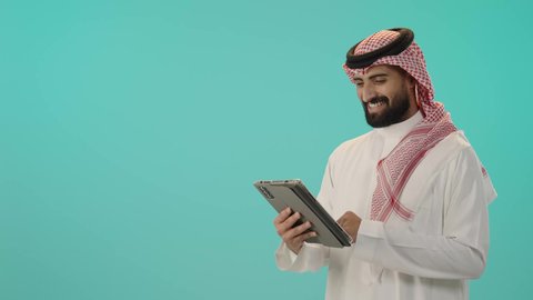 دمج التقنيات الحديثة والأجهزة التكنولوجية بالحياة العملية، بورتريه لرجل عربي خليجي سعودي مبتسم يرتدي الثوب الأبيض التقليدي و الشماغ يمسك جهاز التابلت بيديه، تصفح مواقع الانترنت، الدردشة مع الأهل والأصحاب, خلفية زرقاء