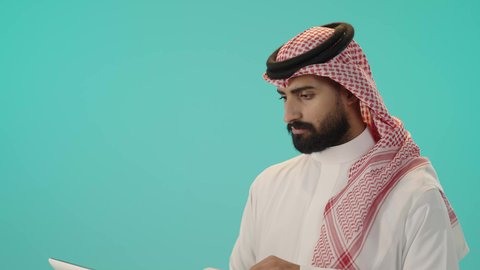 تطور التكنولوجيا والأجهزة التقنية، استخدام جهاز الحاسوب المحمول لإنجاز المهام, تصفح برامج التواصل الاجتماعي, بورتريه لرجل عربي خليجي سعودي يرتدي الثوب التقليدي والشماغ يحمل لابتوب و ينقر على لوحة المفاتيح, ايماءات الاندماج والتركيز, خلفية زرقاء