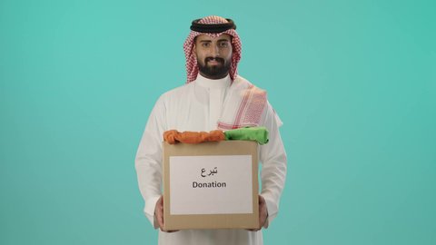 تبرعات ومبادرات انسانية, بورتريه لرجل عربي خليجي سعودي مبتسم يرتدي الثوب الأبيض التقليدي و الشماغ يحمل صندوق كرتوني، النظر إلى الكاميرا بإيماءات السرور، خلفية زرقاء