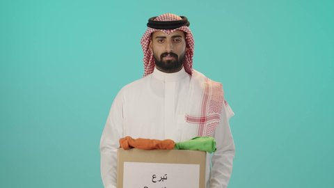 مفهوم التبرع والأعمال الخيرية, بورتريه لرجل عربي خليجي سعودي مبتسم يرتدي الثوب الأبيض التقليدي و الشماغ يحمل صندوق كرتوني، النظر إلى الكاميرا بإيماءات السرور، خلفية زرقاء