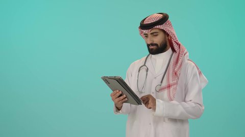 تقديم الاستشارات للمرضى عن بعد عبر الحاسوب اللوحي، بورتريه لطبيب عربي خليجي سعودي يرتدي الثوب التقليدي والشماغ مع المعطف الأبيض يضع سماعة الفحص و يستخدم جهاز التابلت لإعداد التقارير الطبية, دمج التقنيات الحديثة بالمجالات الطبية, خلفية زرقاء