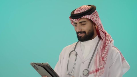 Integrating modern technologies into the medical field, providing remote consultations to patients via tablet computer. Portrait of a Saudi Arabian Gulf Arab doctor wearing a traditional thobe and shemagh with a white coat, wearing a stethoscope and using a tablet to prepare medical reports, Blue background.