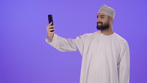 التقاط الصور التذكارية وتوثيق اللحظات السعيدة, بورتريه لرجل عربي خليجي عماني يرتدي الدشداشه والكمه يمسك الهاتف المحمول ويقوم بألتقاط صورة سيلفي لنفسه, مفهوم الأناقة والرجولة والاهتمام بالمظهر الخارجي, خلفية بنفسجية