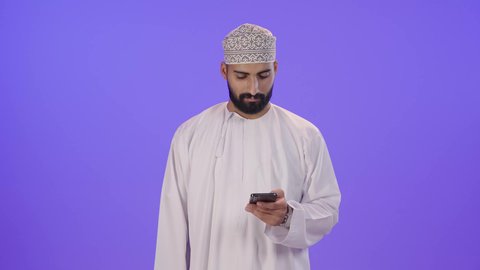 عرض شاشة جوال خضراء، استخدام التقنيات الحديثة والجهزة المتطورة، بورتريه لرجل عربي خليجي عماني يرتدي الدشداشة والكمة يمسك بيده الهاتف المحمول، النظر إلى الكاميرا بإيماءات السعادة والسرور، إعلان ودعاية ترويجية، الجسم بالطول الكامل، خلفية بنفسجية