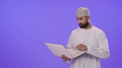 مفهوم ادارة الأعمال عن بعد، استخدام جهاز تقني حديث ومتطور، بورتريه لرجل عربي خليجي عماني يرتدي الدشداشة والكمة يستخدم الحاسوب المحمول، دمج التكنولوجيا بالعمل، إنجاز المهام المطلوبة عبر اللابتوب، خلفية بنفسجية