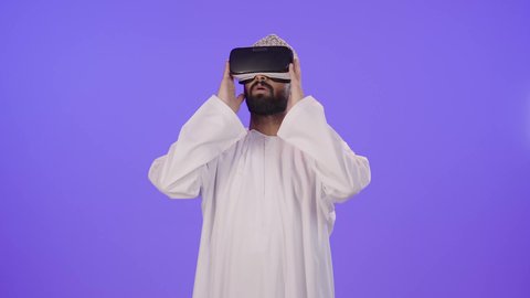 التطور التقني والتكنولوجي، المتعة بالمشاهدة عبر نظارة الواقع الافتراضي, بورتريه لشاب عربي خليجي عماني يرتدي الدشداشة والكمة  ينظر من خلال نظارات ثلاثية الأبعاد (الفي ار), رفع كف اليد للأعلى بإيماءات الاندماج ومحاكاة العالم الخيالي، خلفية بنفسجية