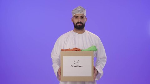 جمعية خيرية ومبادرة انسانية في سلطنة عمان، توفير المستلزمات الضرورية للفقراء، تأمين الملابس وإهدائها للمحتاجين، بورتريه لرجل عربي خليجي عماني يرتدي الدشداشة و الكمة يحمل صندوق من الثياب، مفهوم المساعدة والتبرع، خلفية بنفسجيه
