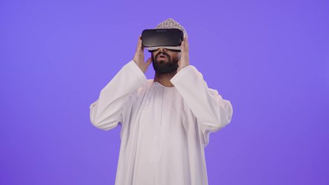 المتعة بالمشاهدة عبر نظارة الواقع الافتراضي، التطور التقني والتكنولوجي, بورتريه لشاب عربي خليجي عماني يرتدي الدشداشة والكمة  ينظر من خلال نظارات ثلاثية الأبعاد (الفي ار), رفع كف اليد للأعلى بإيماءات الاندماج ومحاكاة العالم الخيالي، خلفية بنفسجية