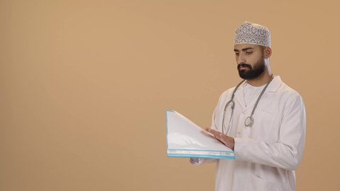 العمل في القطاع الصحي، اعداد التقارير الطبية، تدوين نتائج الفحوصات بالسجل الطبي، بورتريه لطبيب عربي خليجي عماني يرتدي الثوب التقليدي والمعطف الابيض يحمل بيده ملفات المرضى، ايماءات الاندماج و التركيز ، خلفية بني
