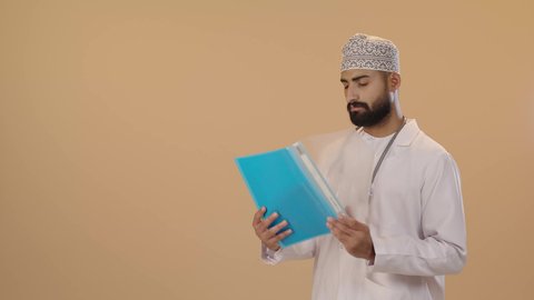 اعداد التقارير الطبية، العمل في القطاع الصحي، تدوين نتائج الفحوصات بالسجل الطبي، بورتريه لطبيب عربي خليجي عماني يرتدي الثوب التقليدي والمعطف الابيض يحمل بيده ملفات المرضى، ايماءات الاندماج و التركيز ، خلفية بني