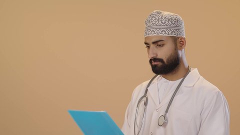 تدوين نتائج الفحوصات بالسجل الطبي، اعداد التقارير الطبية، العمل في القطاع الصحي، بورتريه مقرب لطبيب عربي خليجي عماني يرتدي الثوب التقليدي والمعطف الابيض يحمل بيده ملفات المرضى، ايماءات الاندماج و التركيز ، خلفية بني