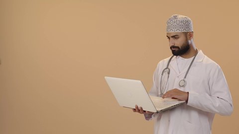 استخدام الأجهزة التقنية الحديثة في الطب، بورتريه لطبيب عربي خليجي عماني يرتدي الدشداشة و الكمة و المعطف الأبيض و السماعات الطبية يحمل بيده الحاسوب المحمول، تقديم الاستشارات الطبية عن طريق الإنترنت، العمل في قطاع الصحة والرعاية، ايماءات الاندماج و التركيز، خلفية بني
