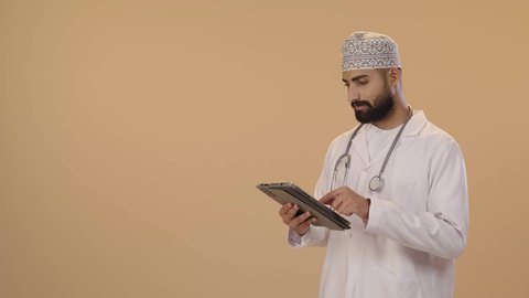 Integrating modern technologies into medical fields, the development of the healthcare sector in the Sultanate of Oman, providing remote consultations for patients through technological devices, a portrait of an Arab Gulf Omani doctor wearing a dishdasha, kumma, and white coat with a stethoscope holding a tablet in his hands, brown background.
