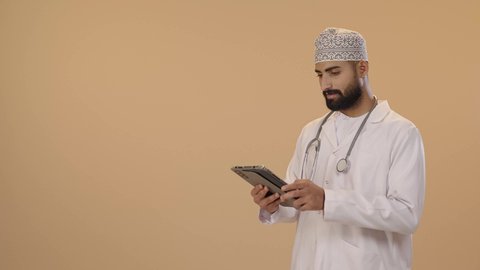تقديم الاستشارات للمرضى عن بعد الأجهزة التكنولوجية، تطور القطاع الصحي في سلطنه عمان، دمج التقنيات الحديثة بالمجالات الطبية، بورتريه لطبيب عربي خليجي عماني يرتدي الدشداشة و الكمة والمعطف الأبيض و السماعات الطبية يمسك جهاز التابلت بيديه، خلفية بنية