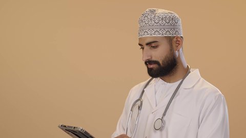 تطور القطاع الصحي في سلطنه عمان، دمج التقنيات الحديثة بالمجالات الطبية، تقديم الاستشارات للمرضى عن بعد الأجهزة التكنولوجية، بورتريه مقرب لطبيب عربي خليجي عماني يرتدي الدشداشة و الكمة والمعطف الأبيض و السماعات الطبية يمسك جهاز التابلت بيديه، خلفية بنية