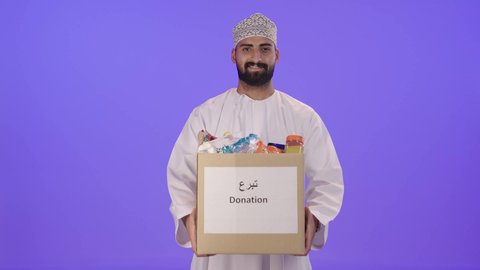 Providing clothing and donating it to those in need, a charitable organization and humanitarian initiative in the Sultanate of Oman, supplying essential items for the poor, a portrait of an Arab Gulf Omani man wearing a dishdasha and kumma carrying a box of clothes, the concept of assistance and donation, purple background.