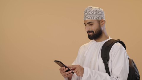 Research and reading through the mobile phone, browsing applications and social media, a close portrait of an Arab Gulf Omani student wearing a dishdasha and kumma and carrying a backpack using a mobile phone, preparing for the new university academic year, brown background.