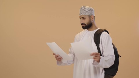 Higher education in Oman, taking notes and writing reports and scientific research, a portrait of an Omani Arab university student wearing a dishdasha and kumma, carrying a backpack and holding a set of papers in his hand, reviewing daily lessons, brown background.