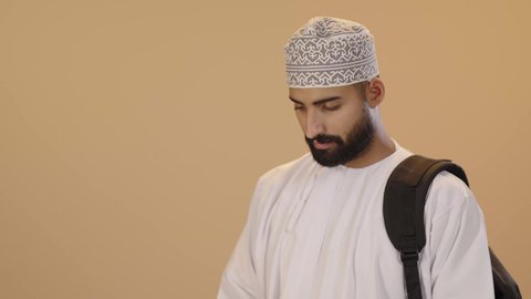 مراجعة الدروس اليومية، بورتريه مقرب طالب جامعي عربي خليجي عماني يرتدي الدشداشة و الكمه يضع الحقيبة على ظهرة يحمل بيده كتاب جامعي، قراءة الكتب الجامعية، التعليم الجامعي في سلطنه عمان ، خلفية بني
